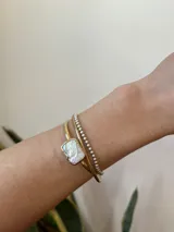 Pulsera rígida dorada con una perla rectangular blanca en el centro.