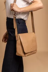 Bandolera de hombro color gris claro, con solapa frontal y correa ajustable. Presenta un diseño simple y práctico, ideal para uso diario.