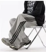 Pantalón deportivo gris claro, estilo cargo o jogging, con franjas negras verticales en los laterales de las piernas. Posee tobillos regulables y cintura ajustable.