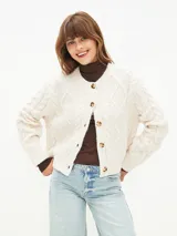 Cardigan de tejido de punto color beige claro, con diseño de trenzas y rombos, cuello redondo y cierre frontal con botones marrones.
