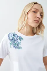Remera blanca de algodón con cuello redondo y mangas cortas. Presenta un bordado de flores paisley en el hombro izquierdo.