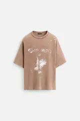 Camiseta color marrón claro con cuello redondo, mangas cortas y estampado frontal con texto e imagen en blanco.