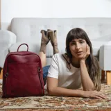 Mochila color borgoña de símil cuero con detalle de costuras en el frente. Tiene un bolsillo frontal con broche magnético, dos bolsillos internos con cierre y un bolsillo abierto con división. Cuenta con asa de mano y correas regulables.