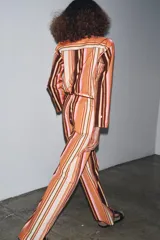 Pantalón de tiro medio con estampado de rayas verticales en tonos marrón, naranja, rosa y beige. Cuenta con trabillas en la cintura, cinturón ajustable con lazadas, bolsillos laterales y cierre frontal con cremallera y botones.