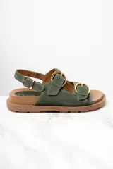 Sandalias de gamuza color verde militar con dos tiras anchas sobre el empeine y una tira trasera, todas ajustables con hebillas doradas. Suela plana color marrón.