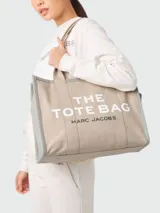 Bolso tipo tote color beige con bordes grises y estampado frontal con el texto "THE TOTE BAG MARC JACOBS" en blanco.