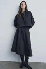 Falda midi negra de gabardina, tiro medio, cintura con trabillas, corte evasé con volumen y cierre frontal con cremallera y botón.