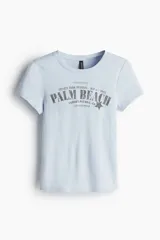 Remera celeste de algodón con cuello redondo y estampado frontal con la frase "Palm Beach Jazz Festival" en letras plateadas con una estrella.
