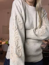 Suéter de tejido de punto color beige claro, con cuello redondo y puños y ruedo acanalados. Presenta apliques florales en relieve con lentejuelas plateadas en el frente y en las mangas.