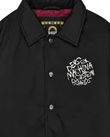 Campera negra tipo coach con cuello clásico, cierre con botones a presión, puños elásticos y cordón ajustable en el ruedo. Estampado en el pecho con el logo de la marca Deus Ex Machina.