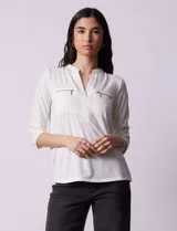 Blusa blanca de poliéster y elastano, con cuello a la base, escote en V y mangas tres cuartos. Presenta dos bolsillos frontales con cierre metálico dorado.