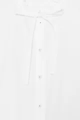 Camisa blanca de popelín con cuello redondo con volante y lazo, manga larga y bajo peplum. Cierre frontal con botones.