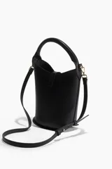 Cartera tipo balde negra de cuero sintético con asa de mano rígida y correa bandolera ajustable y extraíble. Presenta un cierre con trabilla y hebilla metálica dorada.