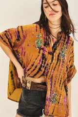 Camisa de manga corta con cuello mao y calce amplio, confeccionada en viscosa con teñido tie-dye en tonos mostaza, negro y violeta. Presenta bordados florales con mostacillas, lentejuelas y canutillos en tonos turquesa y dorado en la parte delantera.