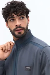 Campera de algodón gris azulado con cierre frontal completo y cuello alto. Presenta detalles de tela técnica azul verdoso en la parte superior del pecho y hombros, y un parche negro con texto blanco en el pecho izquierdo.