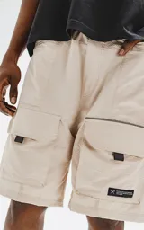 Bermuda color beige con cintura elástica, cordón de ajuste y cinturón negro con hebilla plástica. Tiene bolsillos delanteros y bolsillos cargo con solapa.