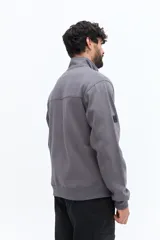 Buzo de algodón gris oscuro con cuello alto y cierre frontal parcial. Presenta un parche de marca en la manga izquierda y un logo bordado en el pecho.
