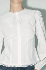 Camisa blanca de popelina de algodón con cuello bobo y mangas largas con elástico en los puños. Detalle de puntilla a tono en cuello y ruedo. Cierre frontal con botones.