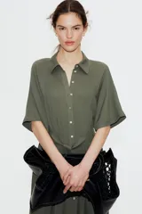 Vestido midi color verde oliva con cuello camisero, botonadura frontal y mangas raglán cortas. Tiene elástico fino en la parte posterior de la cintura, frunces adelante y atrás, y cinturón extraíble para atar.