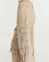 Pantalón cargo de gabardina gastada de lino y algodón color azul. Silueta de pierna recta ancha, con cartera con cierre metálico y broche níquel en el cruce de la cintura. Tiene bolsillos inclinados en los laterales y bolsillos cargo con tapa y broches metálicos a la altura de las rodillas, de los cuales salen unas tiras que se ajustan mediante hebillas metálicas para modificar la silueta del pantalón. En la espalda tiene pinzas y un bolsillo plaque con cierre metálico en boca. Es tiro medio.