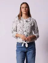 Blusa blanca estampada con flores negras, cuello redondo, abertura con botones y mangas 3/4 con frunce. Tiene terminación con nudo en el ruedo.
