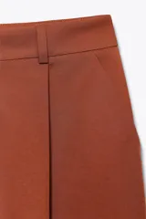 Pantalón marrón de tiro alto, corte ancho y largo midi. Presenta pinzas delanteras, bolsillos laterales y traseros de parche. Cierre frontal con cremallera y gancho metálico.