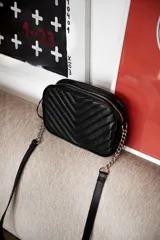 Cartera bandolera negra con diseño capitoneado en forma de V, correa de cuero y cadena plateada, cierre metálico y doble bolsillo interior.