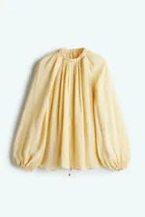 Blusa color crema de gasa transparente con cuello elevado y mangas globo largas con puños elásticos. Presenta pespuntes sobredimensionados decorativos en las costuras raglán y tira diagonal adelante con colgantes de cuentas brillantes.