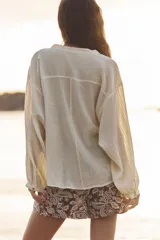 Blusa color crema de tejido arrugado con hilo metalizado, cuello redondo con escote en pico, manga larga abullonada y nudo delantero. Cierre frontal con botones con flores bordadas.