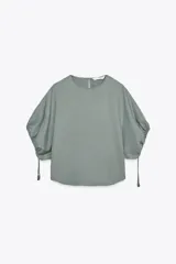 Blusa de cuello redondo color verde, confeccionada en hilatura con mezcla de lyocell. Manga corta abullonada ajustable con lazadas. Bajo redondeado.