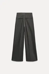 Pantalón de vestir gris oscuro, de tiro alto y corte palazzo. Presenta trabillas, bolsillos laterales y traseros de vivo, pliegues frontales y cierre frontal con cremallera, botón interior y ganchos metálicos.