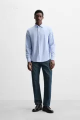 Camisa celeste a rayas verticales blancas, de corte recto, con cuello italiano abierto, manga larga con puños abotonados y cierre frontal con botones.