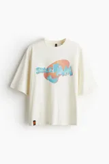 Remera color crema de algodón con estampado del logo de la película Space Jam en tonos celeste y naranja. Corte amplio con cuello redondo y mangas cortas.