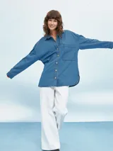 Camisa de denim negro con corte oversize, cuello clásico, cierre frontal con botones metálicos y bolsillo de parche en el pecho.
