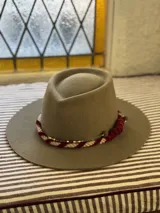 Sombrero de lana color beige con cinta decorativa alrededor de la copa. La cinta combina terciopelo rojo, cordón trenzado blanco y rojo, y detalles de cuentas doradas y marrones.
