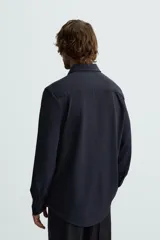 Camisa color beige de corte slim fit, confeccionada en tejido elástico que minimiza las arrugas. Tiene cuello italiano abierto, manga larga con puños abotonados y cierre frontal con botones.