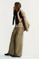 Pantalón color beige de tiro medio con trabillas, bolsillos laterales, de plastrón en pierna y detalle de solapas en espalda. Detalle de pinzas en delantero y cierre frontal con cremallera y ganchos metálicos.