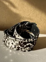 Cinturón de cuero con estampado animal print blanco y negro, tachas redondas y cuadradas plateadas, hebilla metálica y agujeros.