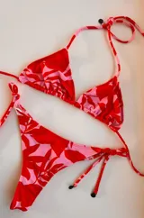Bikini de dos piezas con corpiño triangular ajustable con tiras para atar al cuello y espalda, y bombacha colaless con tiras laterales para atar. Estampado de hojas en tonos rojo y rosa.