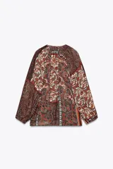 Blusa marrón con estampado de cachemir y detalles de hilo metalizado. Tiene cuello redondo con cordón ajustable y abalorios, escote en pico con nido de abeja, mangas largas con elástico en los puños y cierre frontal con botones forrados.