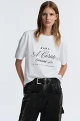 Camiseta marrón de algodón con cuello redondo y mangas cortas. Estampado frontal con el texto "Zara A Coruña Established 1975 A Rúa Juan Flórez 64-66" en color blanco.