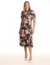 Vestido midi de satén estampado con flores en tonos rosados, blancos y verdes sobre fondo negro. Presenta cuello a la base con frunce, mangas cortas tipo balloon con terminación elastizada, cintura definida y un tajo lateral pronunciado. La espalda cuenta con un lazo para anudar que permite ajustar la caída del cuello. Incluye forro interno a tono.
