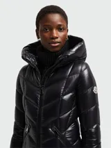 Campera larga de pluma Moncler modelo Marus, color negro con acabado brillante. Presenta un diseño acolchado con costuras en zigzag, capucha integrada, cierre frontal de cremallera y bolsillos laterales con cierre. Incluye el parche con el logo de la marca en la manga.