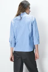 Camisa celeste a rayas verticales blancas, de cuello solapa y manga tres cuartos. Tiene aberturas laterales en el ruedo y cierre frontal con botones ocultos.