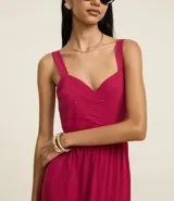 Vestido midi evasê sin mangas color fucsia, confeccionado en tejido airflow con textura arrugada. Presenta escote corazón, breteles medianos con elásticos, recortes fruncidos en la cintura y en el bajo, y cierre lateral con cremallera invisible.