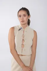 Top tejido color beige con espalda calada y cuello mao. Presenta cierre frontal con cordones y detalle de broches decorativos en el cuello.