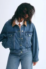Campera de denim azul con cuello de pana marrón, botones metálicos dorados, canesú delantero y trasero, corte holgado, hombros caídos, puños abotonados, bolsillos laterales discretos y bolsillos superiores con solapa y botón.