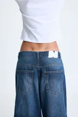 Jean tiro bajo de denim rígido 100% algodón, color azul índigo profundo con matices grises. Presenta un calce recto con pierna amplia y un diseño innovador con corte a la altura de la rodilla, creando un efecto de dos piezas superpuestas con terminación deshilachada.