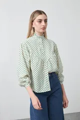 Camisa de viscosa con cuello alto abotonado y mangas largas abullonadas. Presenta un estampado de lunares pequeños en color verde sobre fondo crema.