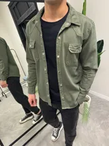 Sobrecamisa verde militar de corte recto, con cuello clásico, cierre frontal con botones metálicos, dos bolsillos con solapa en el pecho y mangas largas con puños abotonados.